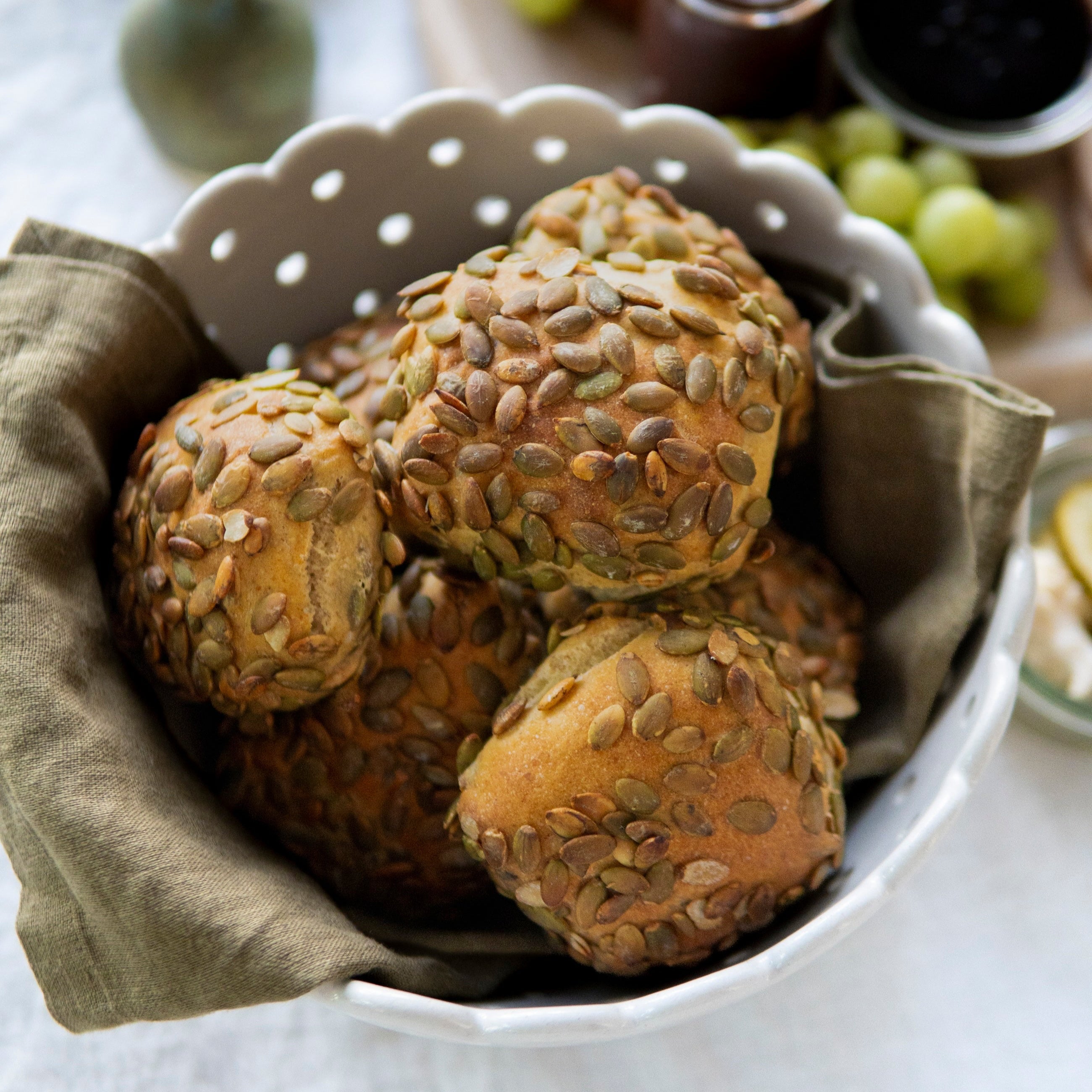 Bowl of seeded bread rolls on a table with fruits and jam in the background